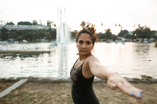 A woman in a black tank top practices yoga by a lakeside fountain at sunset.