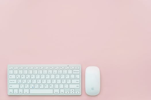 A clean and minimalist flat lay of a white keyboard and mouse on a pastel pink background. Perfect for feminine office themes.