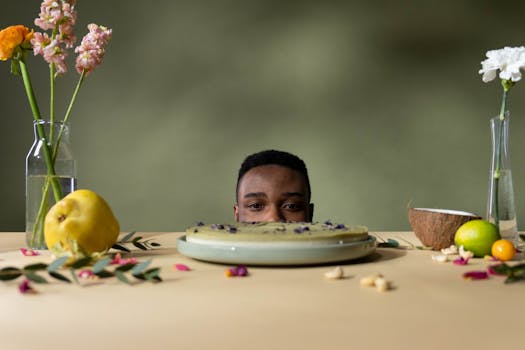 A man peeks over a table with cake, fruits, and flowers, creating a whimsical still life.