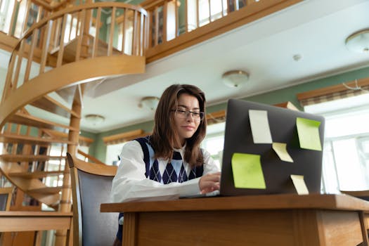 Focused young woman using laptop with notes in a bright room with spiral staircase.