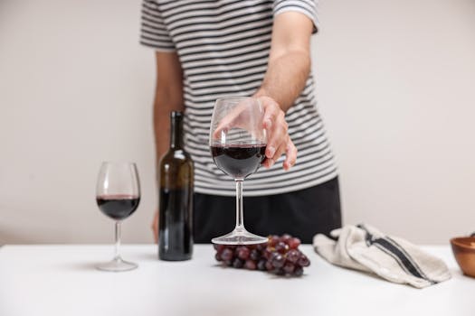 A person in a striped shirt holding a glass of red wine next to grapes and a wine bottle.