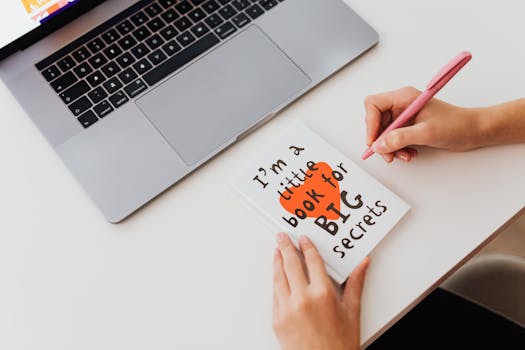 Overhead view of a woman writing in a notebook labeled 'I'm a little book for BIG secrets' beside a laptop.