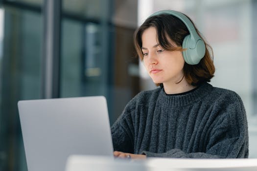 Young woman wearing <a href='https://amzn.to/4mHg7zI' target='_blank'></img>headphones</a> working on a laptop indoors, focused and engaged.