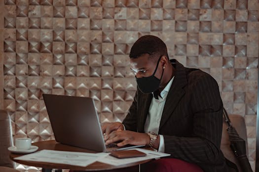 African American man in mask working on laptop in modern indoor setting.