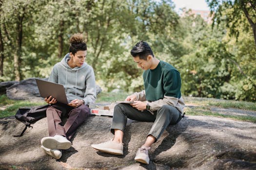 Full body young men in casual clothes spending time on ground with netbook and pizza while taking notes in notebook in summer sunny day with trees on background