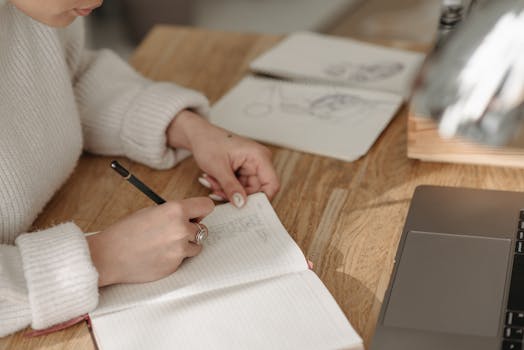A woman writes in a lined notebook at a wooden desk with a laptop and sketchbook nearby.