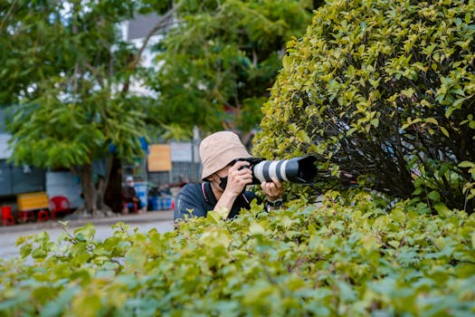 A photographer in a hat captures photos from behind bushes on city streets.