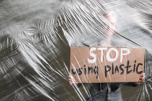 A person holds a cardboard sign reading 'STOP using plastic' behind a plastic sheet, conveying an environmental message.