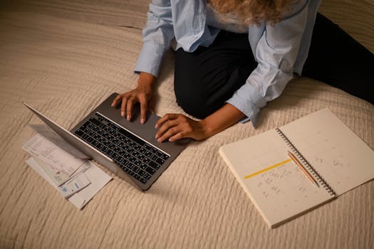 A woman working on a laptop while planning and organizing notes on a bed.