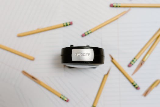 Alarm clock labeled 'Snooze' surrounded by yellow pencils on lined paper, viewed from above.