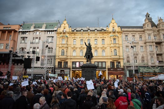 A crowd gathers for a protest at Ban Jelačić Square in Zagreb, Croatia, under a cloudy sky.