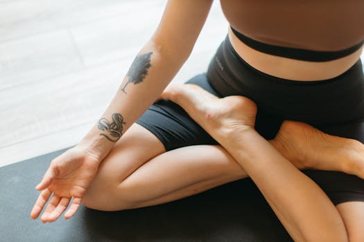 A woman meditates in a yoga pose showing an arm tattoo and flexibility.