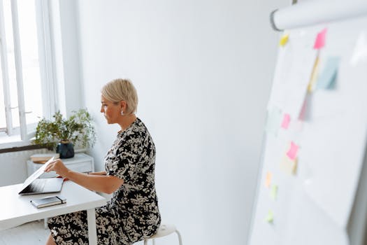 Middle-aged woman working on a laptop in a modern, minimalist office setting with a flip chart.