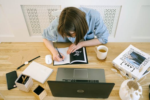 Woman writing notes at a home office desk with a laptop, books, and coffee.