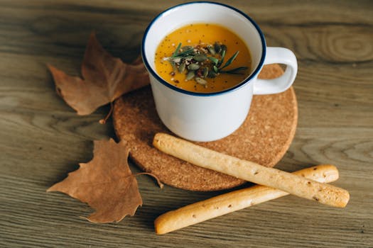 Warm pumpkin soup garnished with seeds and herbs, paired with breadsticks and autumn leaves.