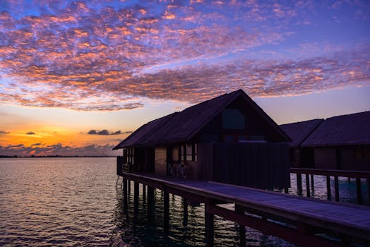 A picturesque overwater bungalow set against a stunning sunset sky with colorful clouds over the calm ocean.