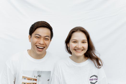 Cheerful couple in white <a href='https://amzn.to/4gUyAb4' target='_blank'></img>t-shirts</a> smiling against a white background, showcasing joy and happiness.