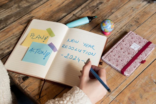 Person writing New Year 2024 resolutions in a notebook with <a href='https://amzn.to/4ntRbNr' target='_blank'></img>sticky notes</a> for planning and action.