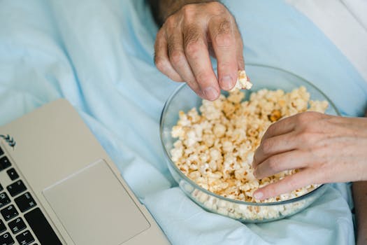 Hands reach for popcorn in a glass bowl next to a laptop, set for a cozy movie night at home.