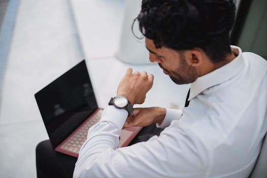 Man in corporate attire looking at his watch while working on a laptop.