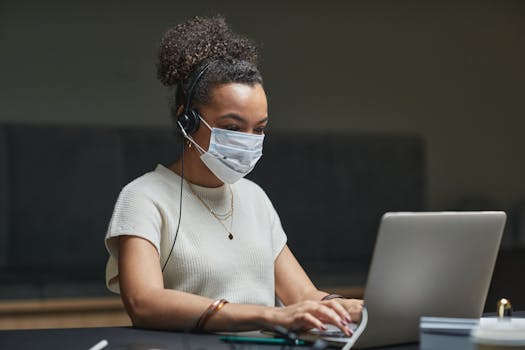 Young woman in a call center setting wearing a mask and using a laptop, demonstrates remote working.