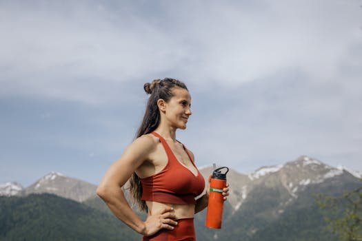 Smiling woman in red sportswear holding a water bottle, standing outdoors against a mountain backdrop.